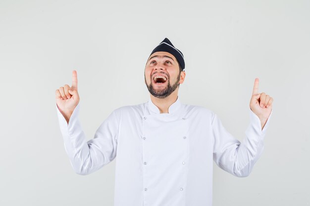 Mannelijke chef-kok die in wit uniform omhoog wijst en er vrolijk uitziet. vooraanzicht.