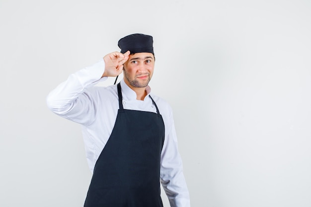 Mannelijke chef-kok die met twee vingers op tempels in uniform, schort groet en er schattig uitziet. vooraanzicht.
