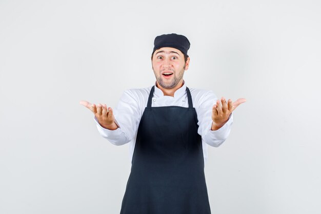 Mannelijke chef-kok die uitnodigt om te komen met handen in uniform, schort en verbaasd. vooraanzicht.