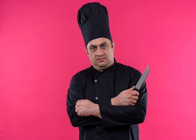Mannelijke chef-kok dragen zwart uniform en kok hoed houden keukenmes met gekruiste handen kijken camera met seriouis gezicht staande op roze achtergrond