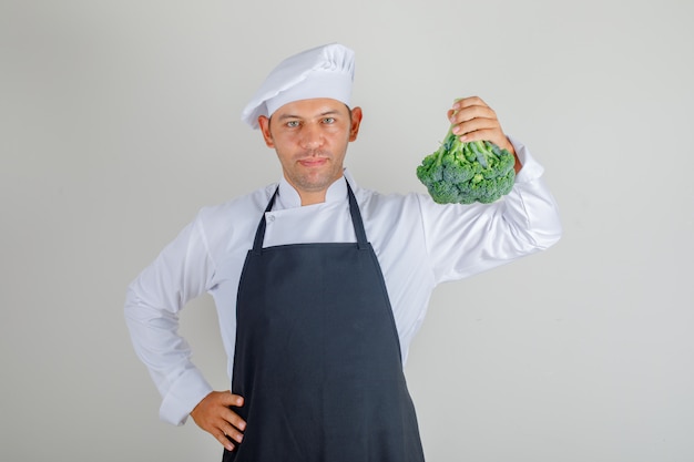 Mannelijke chef-kok in hoed, schort en uniform bedrijf broccoli en hand op taille te zetten