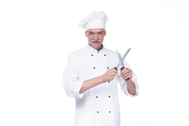 Mannelijke chef-kok in uniform met twee metalen mes terwijl hij op de witte muur blijft