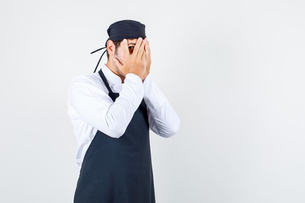 Mannelijke chef-kok in uniform, schort die door vingers met één oog kijkt en verlegen, vooraanzicht kijkt.