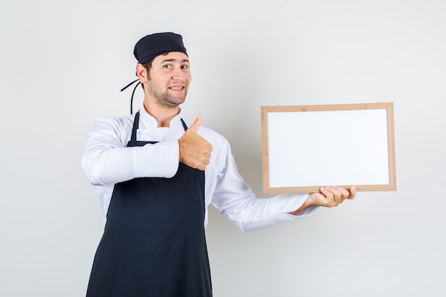 Mannelijke chef-kok in uniform, schort die wit bord met duim omhoog houdt en blij, vooraanzicht kijkt.