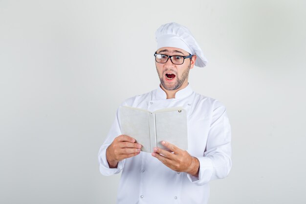 Mannelijke chef-kok in wit uniform, glazen boek lezen en verbaasd kijken