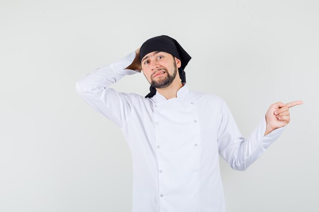 Mannelijke chef-kok in wit uniform wijzend naar de zijkant en besluiteloos kijkend, vooraanzicht.