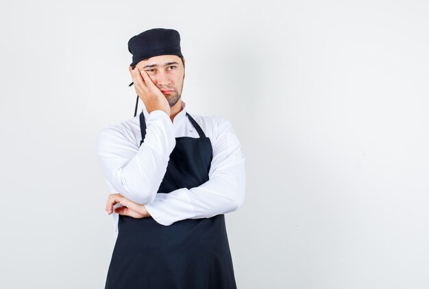 Mannelijke chef-kok leunende wang op opgeheven palm in uniform, schort en peinzend kijken. vooraanzicht.