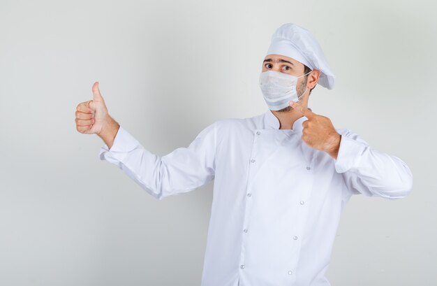 Mannelijke chef-kok medische masker met duim omhoog in wit uniform tonen en voorzichtig kijken