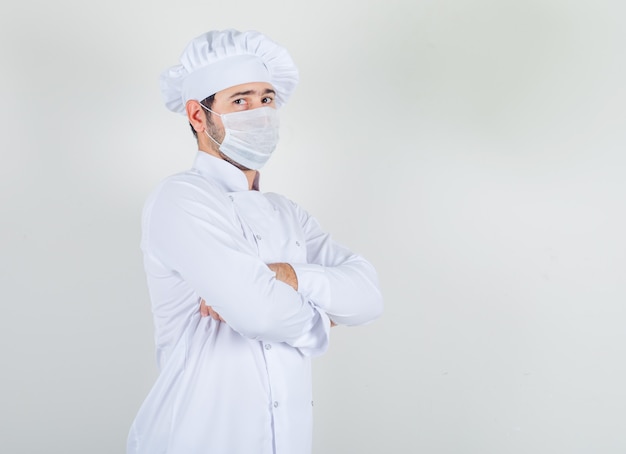 Mannelijke chef-kok permanent met gekruiste armen in wit uniform, medisch masker en voorzichtig kijken