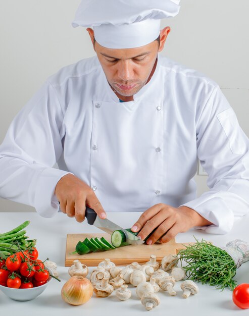 Mannelijke chef-kok zitten en snijden komkommer in keuken in uniform en hoed