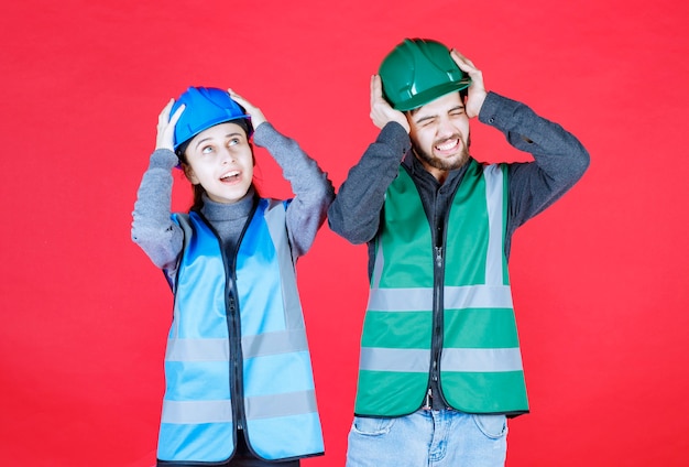 Mannelijke en vrouwelijke ingenieurs die helm en vistuig dragen die het hoofd met twee handen houden.
