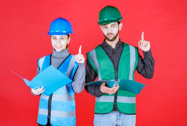 Mannelijke en vrouwelijke ingenieurs met helmen die blauwe mappen vasthouden, deze lezen en mededelingen doen.