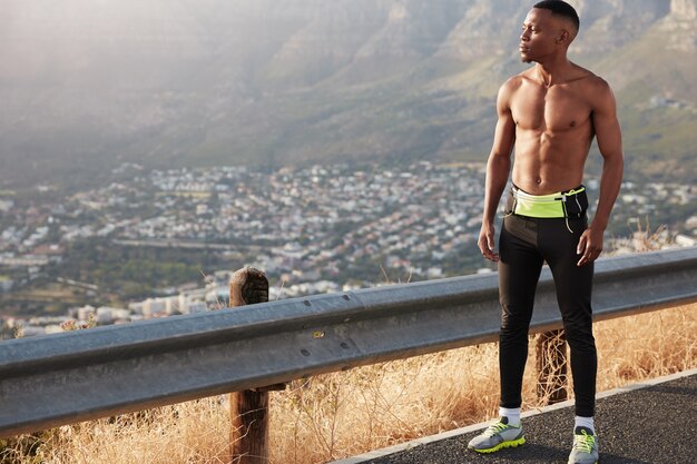 Mannelijke hardloper van volledige lengte met gespierd lichaam, heeft marathon-training, doordachte uitdrukking, bewondert prachtig panoramisch uitzicht op de bergen, denkt na over doeluitdaging, geniet van cardiotraining in de buitenlucht