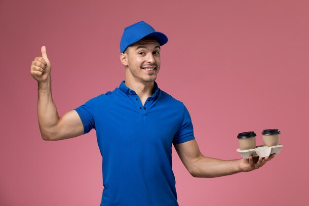 mannelijke koerier in blauw uniform bedrijf koffiekopjes poseren lachend op roze, werknemer uniforme dienstverlening