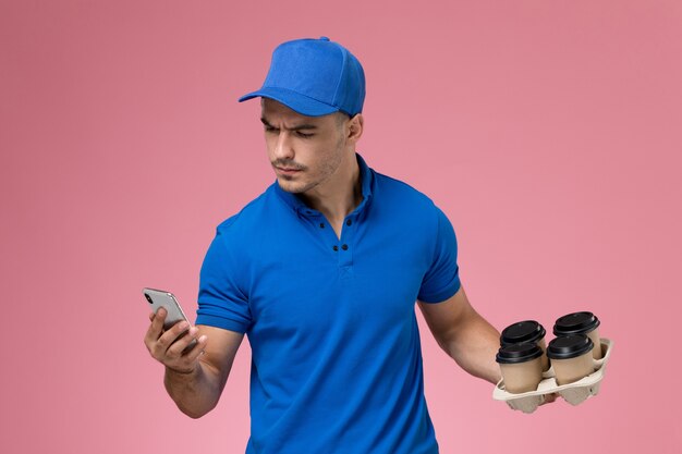 mannelijke koerier in blauw uniform met koffiekopjes met zijn telefoon op roze, uniforme dienstverlening