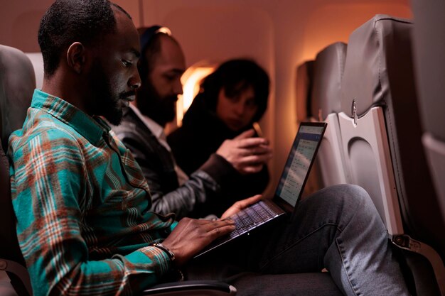 Mannelijke passagier die met het vliegtuig naar het buitenland reist op internationale vlucht, met behulp van luchtvaartdiensten en luchttransport. Freelancer met behulp van laptopcomputer tijdens zonsondergang in economy class.