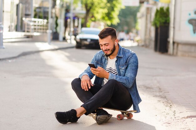 Mannelijke skateboarder zittend op skateboard met behulp van mobiele telefoon