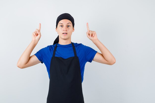 Mannelijke tienerkok wijst omhoog in t-shirt, schort en ziet er zelfverzekerd uit. vooraanzicht.