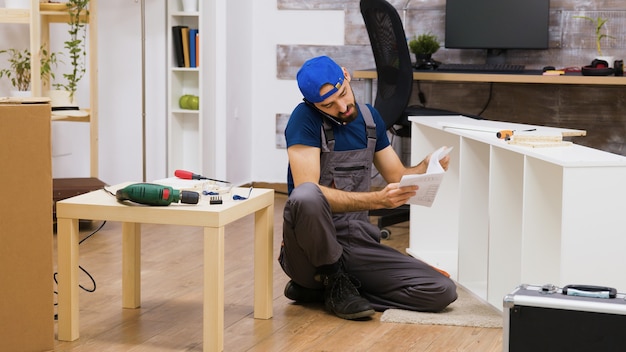Mannelijke werknemer die aan de telefoon praat terwijl hij witte meubels in een nieuw huis monteert. elektrische boor.