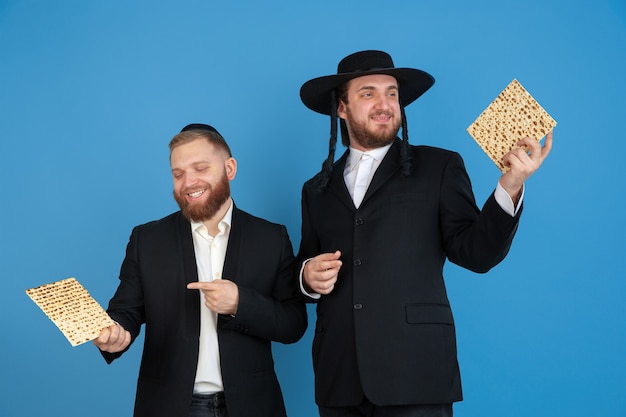 Gratis foto matzah eten, uitnodigend. portret van een jonge orthodoxe joodse mannen geïsoleerd op blauwe muur. purim, zaken, festival, vakantie, viering pesach of pesach, jodendom, religie concept.