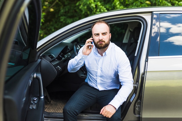 Medio volwassen mensenzitting in een auto met open deur die op smartphone spreken