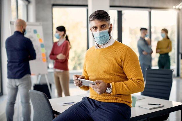 Medio volwassen zakenman die aan touchpad werkt en een beschermend gezichtsmasker op kantoor draagt