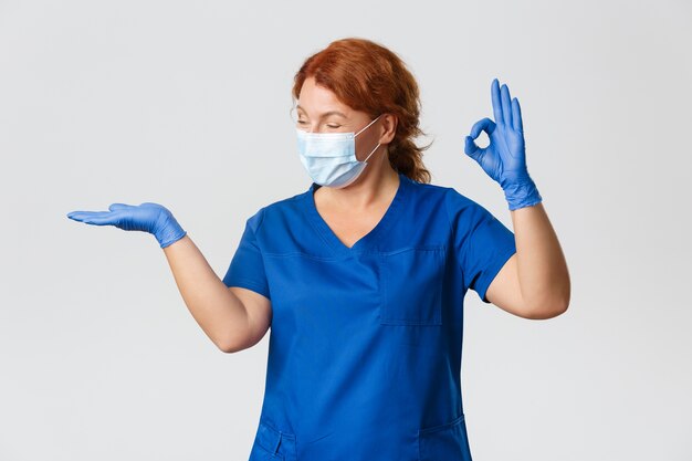 Medisch personeel, pandemie, coronavirus concept. Gelukkig glimlachende vrouwelijke arts, dierenarts of arts in gezichtsmasker en handschoenen, houdt iets op de handpalm en laat zien dat het goed is, aanbevelen