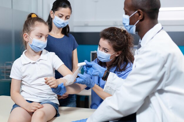 Medisch verpleegkundige helpt afro-amerikaanse kinderarts arts om gebroken arm van kleine jongen patiënt te verbinden tijdens klinische fysiotherapie in ziekenhuis kantoor. Team met gezichtsmasker tegen covid19