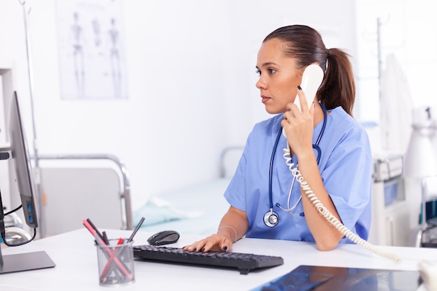 Medisch verpleegkundige in gesprek met patiënt aan de telefoon over diagnose. Gezondheidszorg arts zit aan bureau met behulp van computer in moderne kliniek kijken naar monitor.