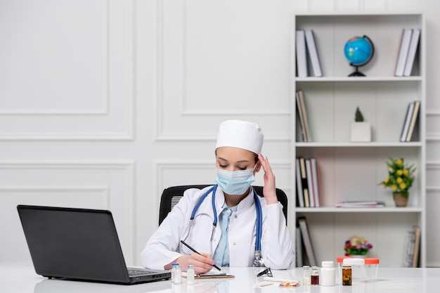 Medische jonge blonde dokter in witte laboratoriumjurk en hoed met computer
