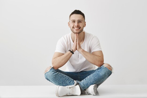 Meditatie en emoties concept Aantrekkelijke stijlvolle jonge man met baard met bril en trendy outfit zittend met gekruiste handen op de vloer terwijl hand in hand bidden en glimlachen over grijze muur