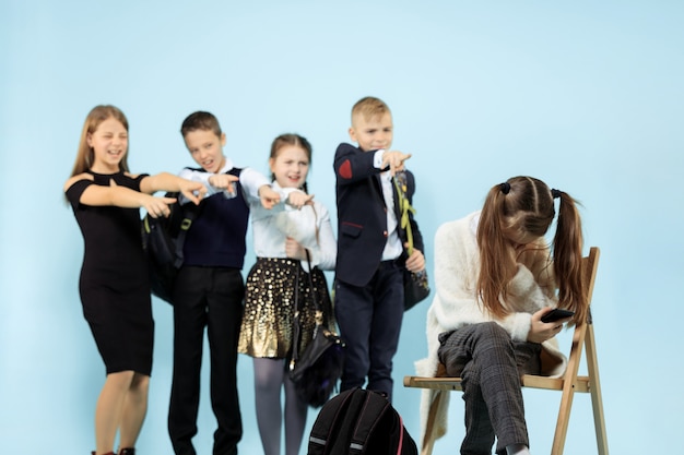 Meisje alleen zittend op een stoel en lijden aan een daad van pesten terwijl kinderen spottend. Triest jong schoolmeisje zittend tegen blauwe muur.