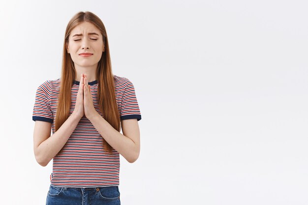 Meisje bidt god, heb geloofsdromen die uitkomen als je gelooft. Aantrekkelijke blonde vrouw in gestreepte t-shirt gesp handpalmen hand in hand in bidden, ogen sluiten en nerveus glimlachen, staande witte achtergrond