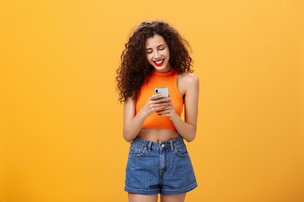 Meisje brengt tijd door op internet en sms't vriendberichten via smartphone terwijl ze lacht terwijl ze naar het scherm van het apparaat kijkt dat blij en vrolijk staat over oranje achtergrond in bijgesneden top en denim shorts.