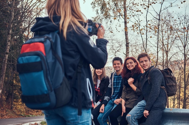 Meisje dat een foto maakt van haar vrienden. Groep jonge vrienden die op vangrail dichtbij weg zitten. Reizen, wandelen, avontuur concept.