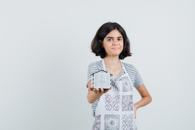 Meisje dat huismodel in t-shirt, schort voorstelt en vrolijk kijkt.