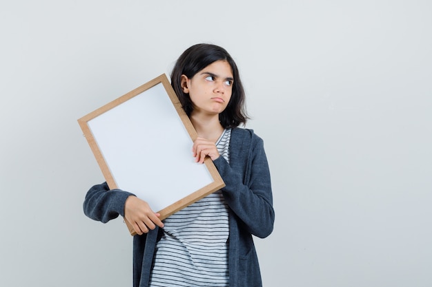 Meisje dat leeg frame in t-shirt, jasje houdt en peinzend kijkt.