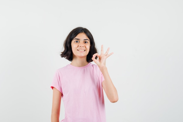 Meisje dat ok gebaar in roze t-shirt toont en joviaal kijkt. vooraanzicht.