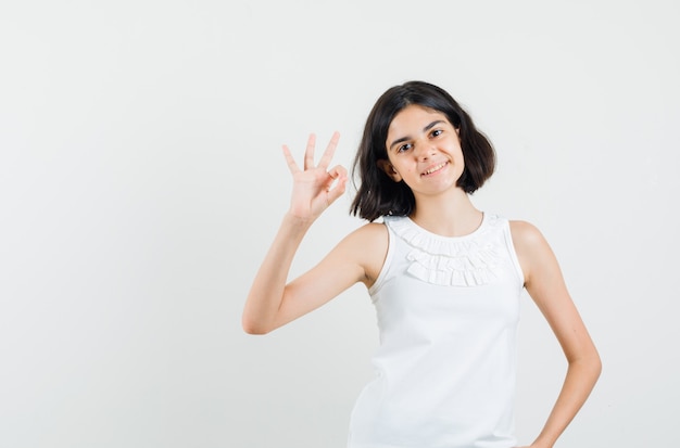 Meisje dat ok gebaar in witte blouse toont en tevreden, vooraanzicht kijkt.