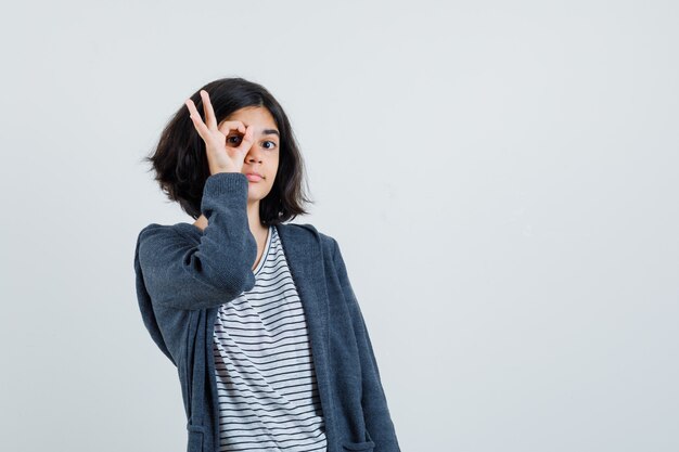 Meisje dat ok teken op oog in t-shirt, jasje toont en nieuwsgierig kijkt