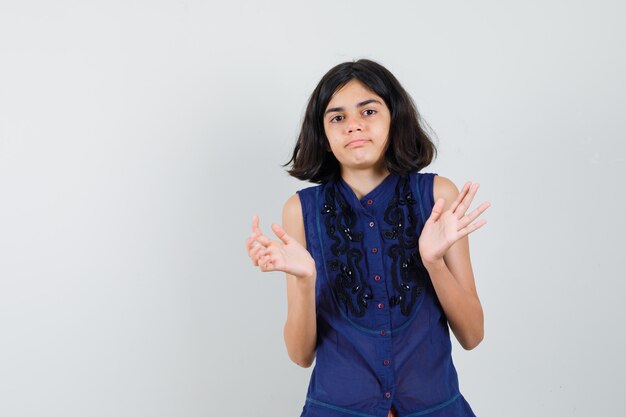 Meisje dat open handpalmen in blauwe blouse opheft en verward kijkt.