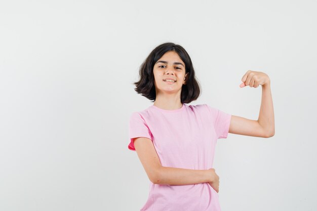 Meisje dat spieren van de arm in roze t-shirt toont en zelfverzekerd, vooraanzicht kijkt.