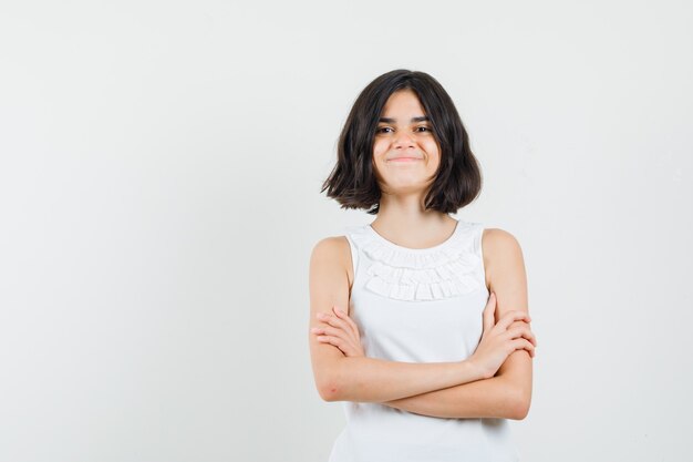 Meisje dat zich met gekruiste wapens in witte blouse bevindt en joviaal kijkt. vooraanzicht.