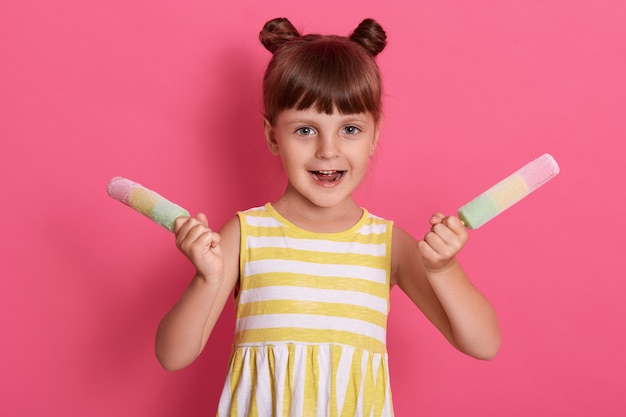 Meisje die kleurrijke ijslollies houden, gezichtsuitdrukking hebben opgewekt, die met gelukkige blik stellen, die witte en gele gestripte de zomerkleding dragen tegen roze muur.