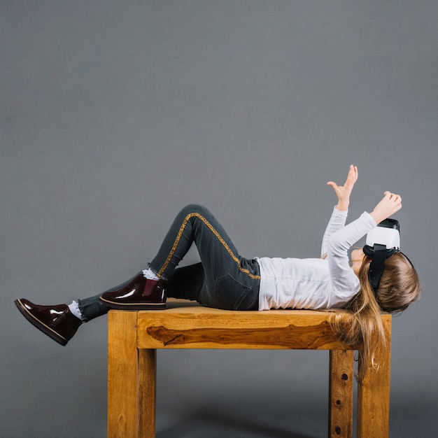 Meisje die op houten lijst liggen die virtuele werkelijkheidsglazen dragen die handgebaar maken tegen grijze achtergrond