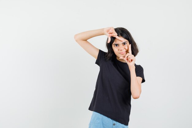 Meisje frame gebaar maken in zwart t-shirt, korte broek en op zoek gericht. vooraanzicht.