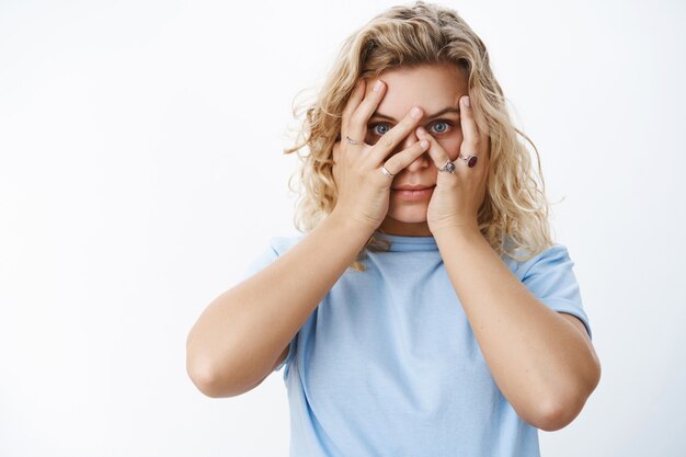 Meisje gluurt omdat ze niet van verrassingen houdt. Leuke en jonge aantrekkelijke blonde met kort kapsel in blauw t-shirt hand in hand op gezicht, zicht bedekken en door vingers gluren om te zien, ongeduldig over witte muur