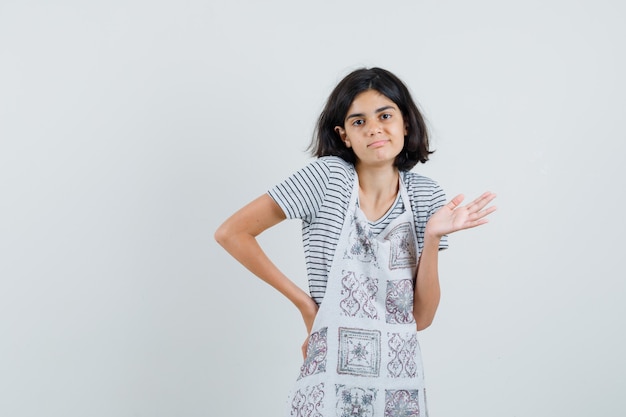 Meisje haalt haar schouders op in t-shirt, schort en kijkt verward.