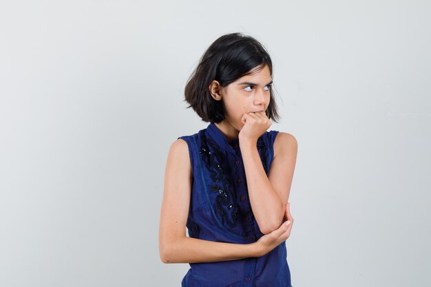 Meisje haar vuist in blauwe blouse bijten en vergeetachtig op zoek. vooraanzicht.