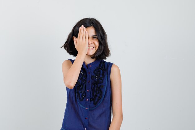 Meisje hand op één oog in blauwe blouse en op zoek vrolijk, vooraanzicht.